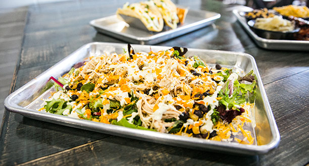 A metal tray with fully loaded nachos sits on a table next to a tray with tacos and other fare.